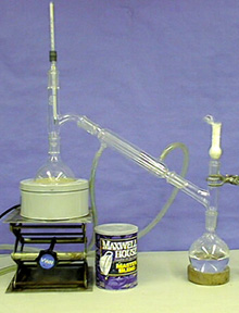 A set up showing a round bottom flask with a clear liquid and an upper white solid attached to a glass tube. The glass tube is connected to a second flask atop a stand with glass tubing.