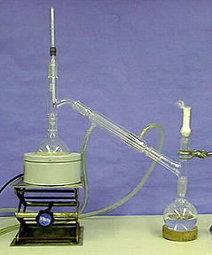 A set up showing a round bottom flask with a clear liquid and an upper white solid attached to a glass tube. The glass tube is connected to a second flask atop a stand with glass tubing.