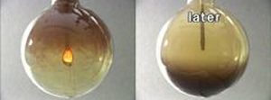 A metal spoon in a flask with fire, dark brown smoke in the upper layer and a yellow gas the lower layer. A metal spoon with a yellow-brown upper layer and a dark brown lower layer in a large flask.