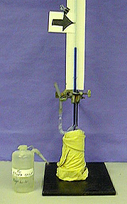 A glass tube with blue liquid on a ring stand with white background, an arrow, and a yellow cloth wrapped glass container. A squirt bottle with a label beside the ring stand.