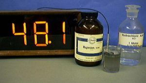 A thermometer, a capped, dark bottle labeled Magnesium, a cable and temperature probe in a beaker with cloudy liquid, and a capped bottle with clear liquid labeled Hydrochloric Acid