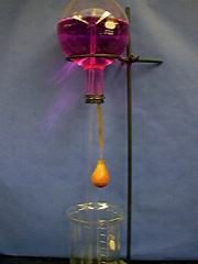 An inverted round bottom flask with red liquid, glass tubing, and silicone tubing with an orange bulb. A ring stand holds the  flask above a beaker.