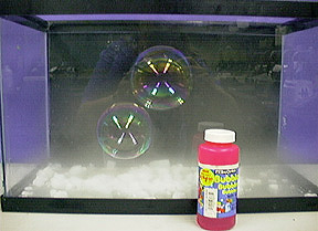 Two bubbles float inside an aquarium tank. Vapor and white solids are at the bottom of the aquarium tank. A capped pink bottle labeled "bubbles" is in front of the tank with a purple background.