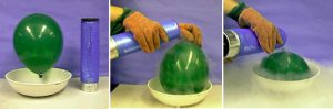 In the first photo, an inflated balloon is suspended in a white bowl with a black capped container. In the second photo, a gloved hand pours the container's contents over the green balloon in the white bowl. In the third photo, a gloved hand pour clear liquid from the blue container on to the shriveled balloon while vapor surrounds the balloon in a bowl.