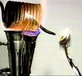 A copper colored coil with a blackened tube over two burners, white sparks, and a solid held by tongs