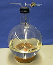 A round flask with a two-hole stopper and glass tubes contains clear liquid and orange boiling chips. The flask is labeled "acetone" and sits on a cork ring.