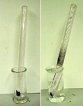 The first photo shows a glass tube with a black substance at the bottom of the tube, and the tube is in an empty clear glass container. The second photo shows a glass tube with a black substance at the bottom of the tube and dark specks in the middle and upper portions of the tube. The tube is in clear liquid inside a glass container.