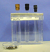Three corked test tubes with clear liquids in a rack with identifying labels of the liquids. Metal silver and black balls are in a clear dish in front of the rack.