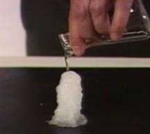 A hand pours clear liquid from a flask onto a tower-shaped white solid on a dark surface.