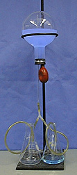 An inverted round bottom, stoppered flask with fluorescent blue liquid is connected to an orange bulb and glass tube attached to a Y-connector. The Y-connector is attached to two tubes, with each end of the tube placed in a flask. The flask on the right contains more clear liquid than the left flask contains. The inverted flask is held by a ring stand.