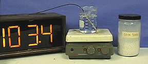 A thermometer readout higher than the boiling point of water, a temperature probe with an attached cable in a beaker with clear liquid atop a hot plate, and a capped jar with white solid with a yellow label
