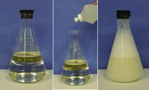 The first photo shows a stoppered flask with an upper layer of yellow liquid and a lower layer of clear liquid that reflects the yellow liquid. In the second photo, a plastic bottle nozzle is held above the unstoppered flask with the upper layer of yellow liquid and the lower layer that reflects the yellow liquid. The third photo shows the stoppered flask with bubbles in the uppermost layer, white foam in the middle layer, and a darker foam in the bottom layer.