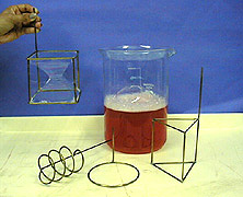 A hand holds a square shaped bubble wand with soap film. Various geometric shaped wands and a beaker containing an upper layer of foam and a lower layer of red liquid soap appear in the photo.