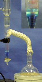 A trap containing a blue liquid is connected to a stop cock and wrapped tubing, and the tubing is attached to a flask in a  white support container. The insert shows a close-up photo of layered blue and clear liquids inside the nozzle.