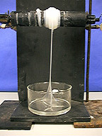 White thread-like substance is pulled from a container with a clear mixture and wound around a windlass. The container sits atop a black base and a black background.