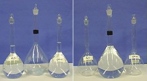 Two photos, each with three stoppered flasks. The first photo shows a labeled flask with clear liquid in front of  a blue background, an empty flask with a black band, a labeled flask with another clear liquid. In the second photo, the flask with the black band has cloudy liquid. The remaining two flasks are empty.