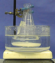 A ring stand with clamp holds in place a flask with clear liquid and a white solid in the bottom of the flask. The flask is held in a container with clear liquid atop a white surface.