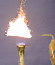 A white flame with orange sparks appears over a blue flame of a burner. A squirt bottle is shown next to the flame and burner spraying iron powder into the burner flame.
