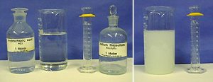 In the first photo, a stoppered, labeled bottle with clear liquid, a beaker with clear liquid, a graduated cylinder with a yellow bumper, and a stoppered, labeled bottle with a clear liquid.  In the second photo, a beaker with a pale precipitate and a graduated cylinder with a yellow bumper.