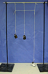 Three weights hanging from strings tied together, two black and one wrapped in white paper, hang from a crossbar. The crossbar is supported by two stands with black bases.