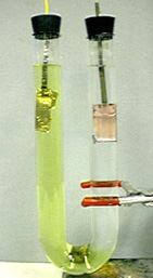 A U-tube with a two-colored solution, clear yellow and clear. Two electrodes in the tube are shown, one is silver colored and one coppered colored with a silver bottom.