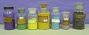 One capped, labeled bottle and six stoppered, labeled bottles, each with solids. The colors appear respectively: blue, green, yellow, brown-yellow, light blue, salmon, and dark gray.