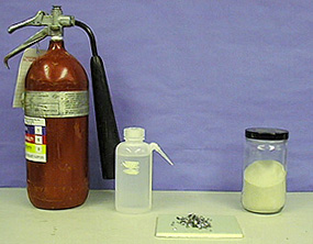 A fire extinguisher with a nozzle, a squirt bottle with clear liquid, silver-colored shards on a white surface, and a capped jar with yellow sand