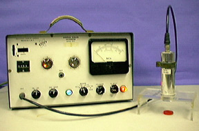 An instrument used to measure radiation with a row of dials, a probe attached to the instrument, and a silver cylinder connected to a plexiglass stand. A red disc is seen beneath the plexiglass stand.