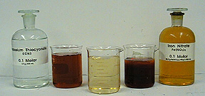 A photo shows a capped, labeled bottle with clear liquid, a beaker with red liquid, a beaker with yellow liquid, a beaker with dark red liquid, and a stoppered, labeled bottle with orange liquid.
