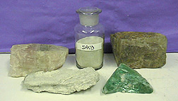 A light brown solid, a gray-white solid, green glass, a brown solid, and a capped bottle labeled "sand" with a pale solid arranged against a blue background