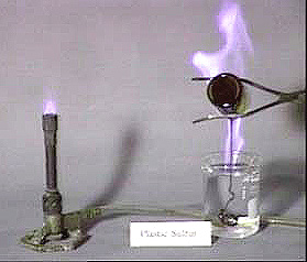 A lit burner connected to a clear silicone tube. Tongs pouring a burning black liquid sulfur into a beaker of clear liquid. The label "Plastic Sulfur" appears at the bottom of the photo.