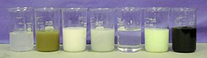 Seven beakers: A beaker with cloudy, silver liquid, a beaker with brown solid, a beaker with white solid, a beaker with gray-white solid, a beaker with clear liquid, a beaker with pale yellow solid, and a beaker with black solid