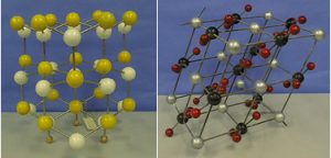 Two ball and stick models representing molecules. The model on the left uses yellow and white spheres and silver rods. The model on the right uses silver, red, and black spheres and silver rods.