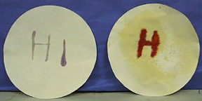 A pair of dark colored letters appear on filter paper. In the second photo, a red letter H is on a yellow filter paper.