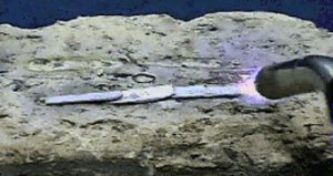 Three silver colored metal strips rest on the surface of a brick. A lit torch with orange-white flame is seen next to the bar on the right.