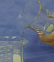 A hand holds a beaker with clear liquid, and the liquid flows from the upper beaker into the lower beaker.