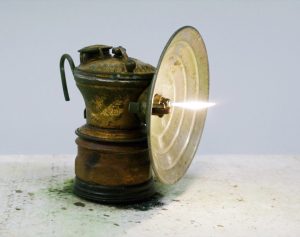 A brass lamp emitting white flame sits on a pale surface against a gray background.