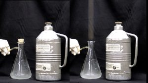 The first photo shows a gloved hand holding tubing connected to a stoppered sidearm flask with cloudy liquid next to a capped metal tank. In the second photo, the gloved hand holds the tube connected to the unstoppered flask with cloudy liquid, and vapor rises from the unstoppered flask.