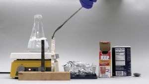 A flask with bubbling liquid sits atop a hot plate. A gloved hand uses tongs to hold an untarnished strip next to a tarnished strip on a wooden block. A pan of aluminum foil and a carton of salt flank a box of baking soda.