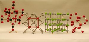 Four models using spheres and rods to represent crystals. The model on the left uses red and black spheres, and silver colored rods. The second model uses gold and red spheres, and gold and red rods. The third model uses green and gold spheres, and dark rods. The model on the right uses large red spheres and smaller white spheres, and dark rods.  
