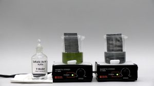 A capped bottle labeled "Sulfuric Acid" on a white cloth, a dark metal strip in yellow liquid inside a beaker atop a stirrer, and a dark metal strip in dark liquid inside a beaker atop a stirrer. A white stir bar is seen in each beaker.