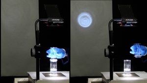 In the first photo, a gloved hand holds a glass cylinder of clear liquid above a second cylinder with liquid on a projector deck. No light is seen on the screen. In the second photo, the hand moves the container away from the container with liquid, and a blue image of the container with liquid is seen.