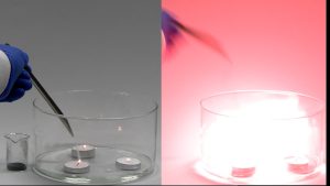 In the first photo, a hand holds a scoopula with powder over three burning candles in a dish with water. A beaker with a black solid is seen next to the dish. In the second photo, red flame is seen inside the dish, and the shadow of the nozzle is seen.