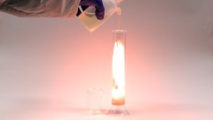 A gloved hand squirts a yellow liquid into a column with an orange-white flame. A liquid is seen at the bottom of the column.