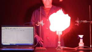 A computer screen used to show wavelengths, a reflector clamped on a ring stand, a red flame atop a burner with a cable, a squirt bottle with liquid, and a fiber optic detector clamped on a stand. The demonstrator is seen in the photo.
