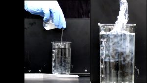 A gloved hand pours a cloudy liquid into a beaker containing a clear liquid. A film-like substance appears in the liquid in the beaker. The second photo shows the substance below the water line in this beaker and a semi-solid mass that has been lifted out above the water line in the beaker.