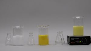 A flask next to a beaker with cloudy white liquid, a flask next to a beaker with cloudy orange liquid, and two flasks, next to a beaker with yellow-white liquid atop a hot plate.