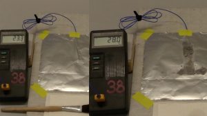 A two photo sequence: In the first photo, a thermometer, a cable taped to aluminum foil and the aluminum foil taped to a surface, and a paintbrush. In the second photo, the thermometer with a different readout with cable taped to aluminum foil, and a darker area on the aluminum foil.