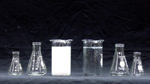 A photo shows two pair of flasks, larger and smaller flanking a beaker with white precipitate atop a gray surface next to a second beaker containing clear liquid with bubbles.