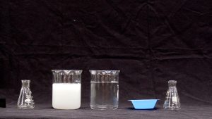 Two small flasks flanking two beakers, one with a white solid. One with clear liquid next to a blue weigh boat.